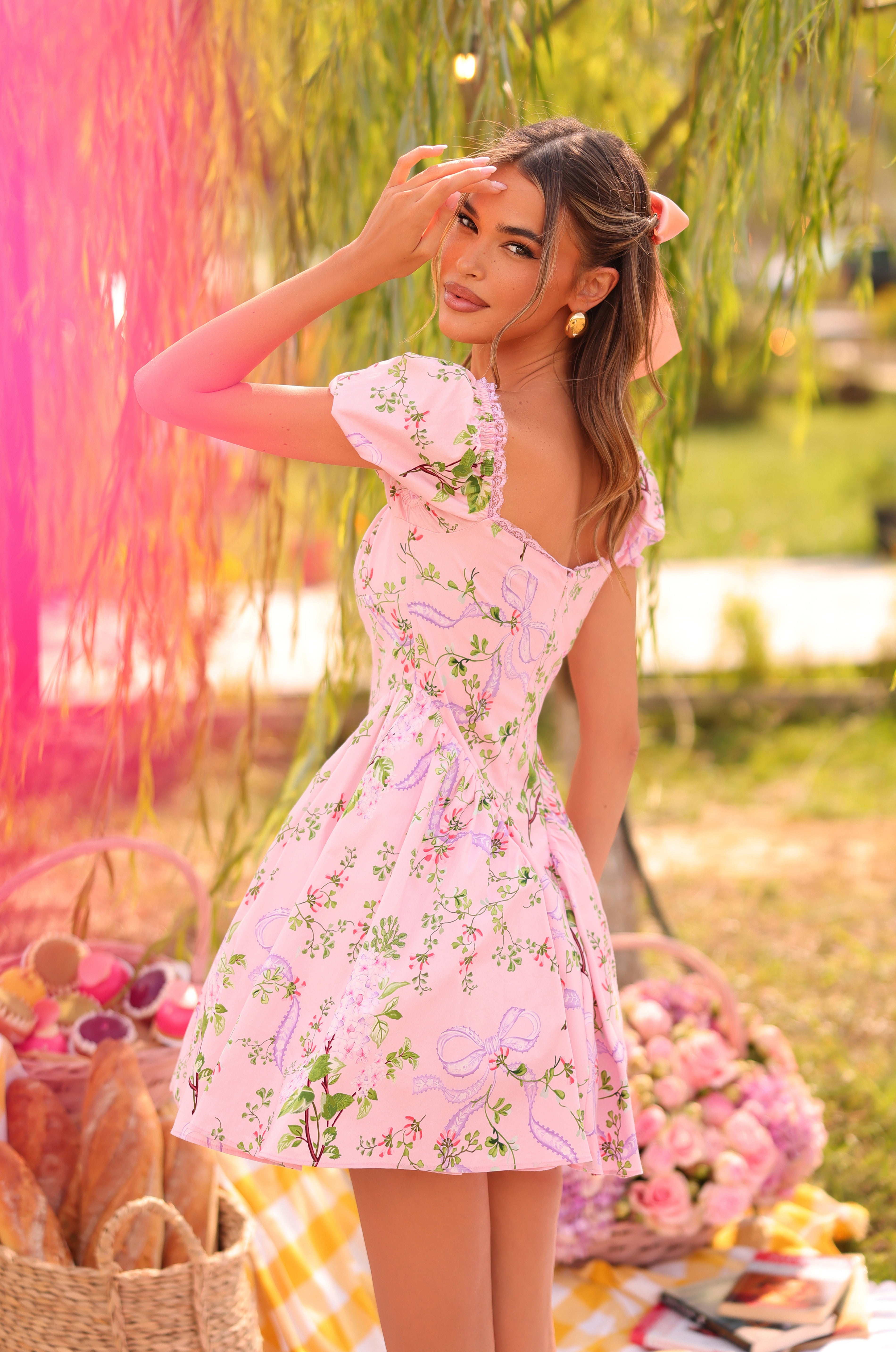 A woman wearing the Blush Bloom Floral Short Casual Dress: Rosabella stands by a picnic setup with baskets, bread, and flowers, amid lush greenery outdoors.